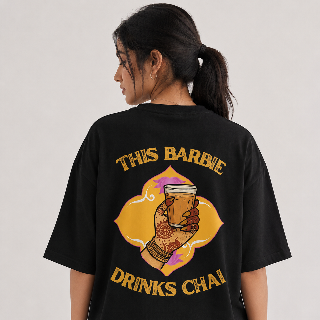 A woman shown from the back wearing a black oversized graphic T-shirt. The shirt features a large yellow and orange illustration of a hand holding a cup, with the text 'THIS BARBIE DRINKS CHAI' in yellow.